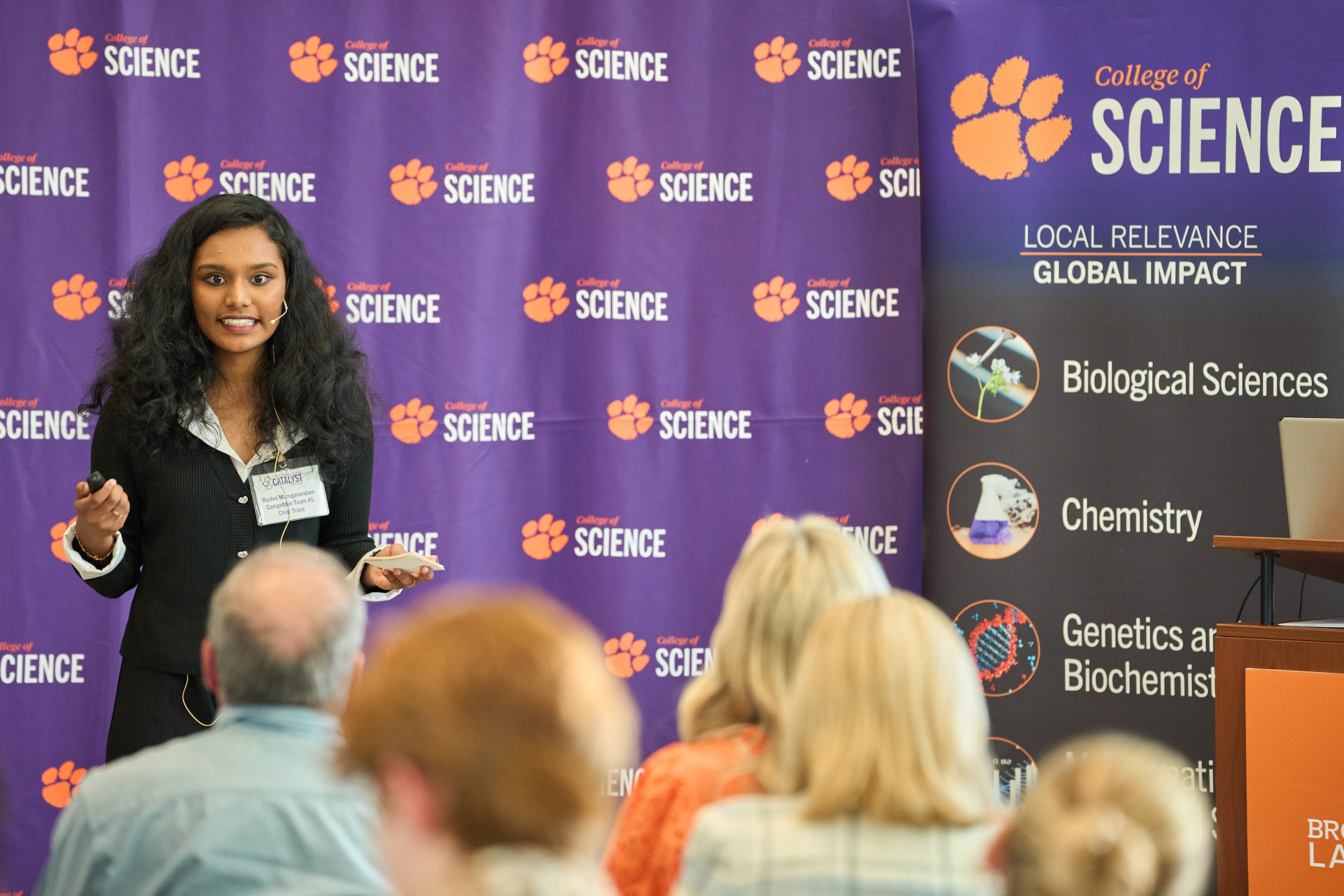 Woman presenting at a conference in front of step-and-repeat background, with College of Science and tiger paw logos.