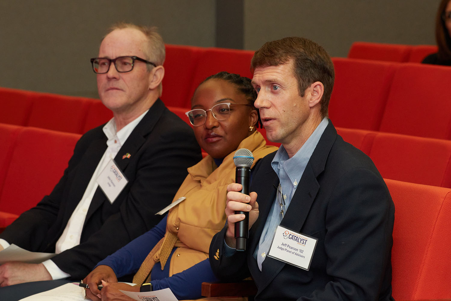 Man, woman, man with microphone sitting in orange seats in a theater-type setting.