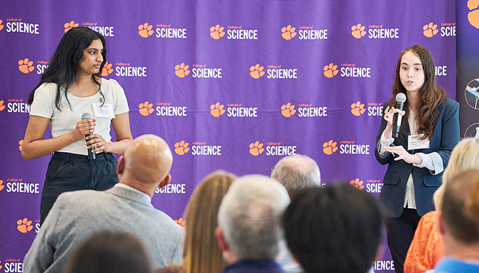 Two people presenting in front of a purple step-and-repeat wall with College of Science logo with tiger paw.