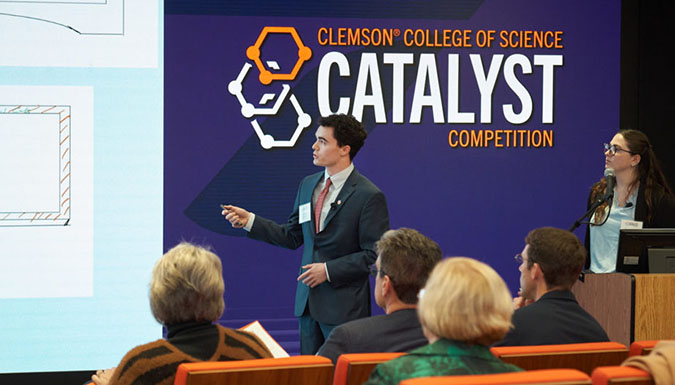 Student presents his project to an audience. Catalyst logo on screen behind him, while female student looks on from podium.
