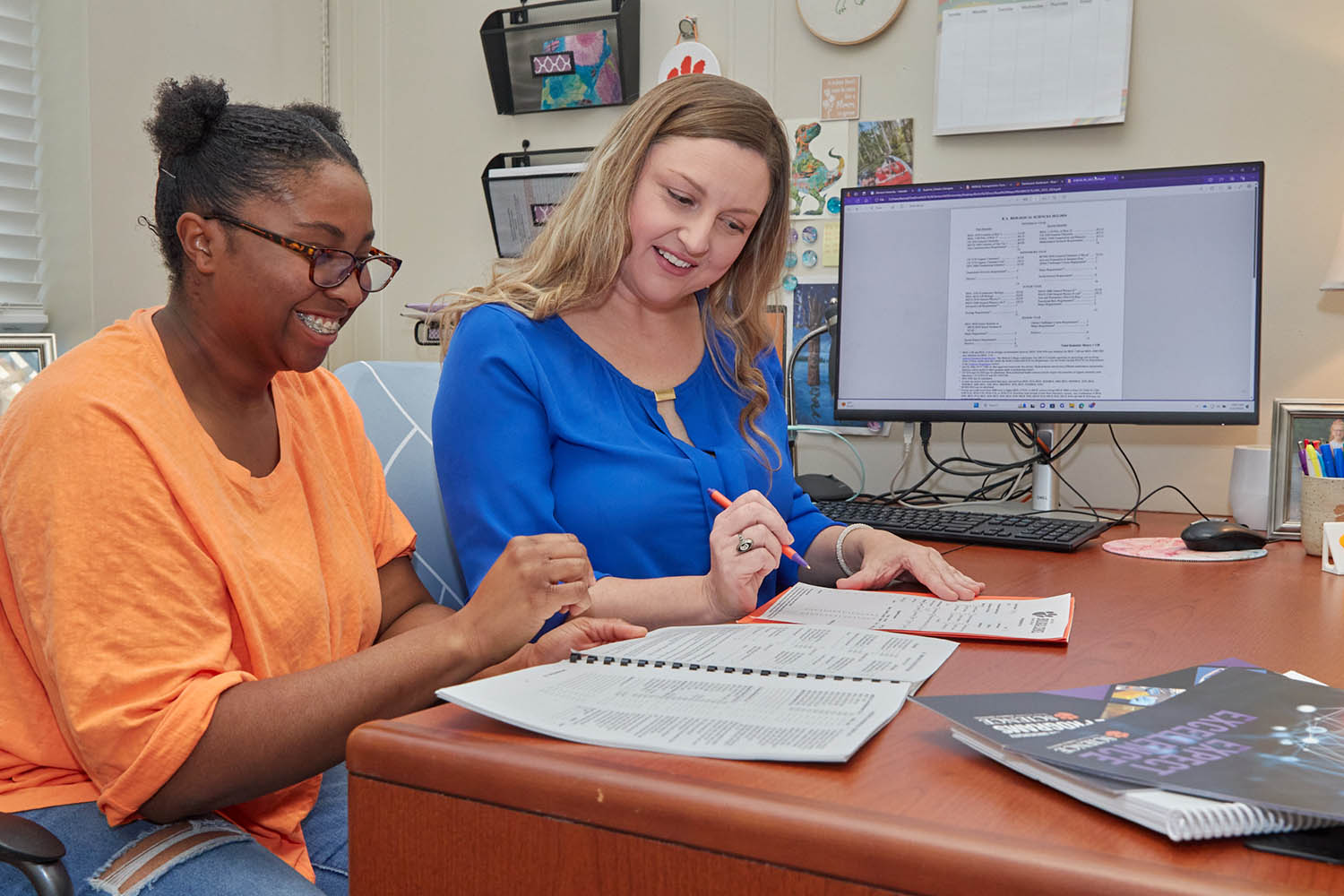 Two female students in a classroom setting, one wearing a pink top, one wearing a gray top.