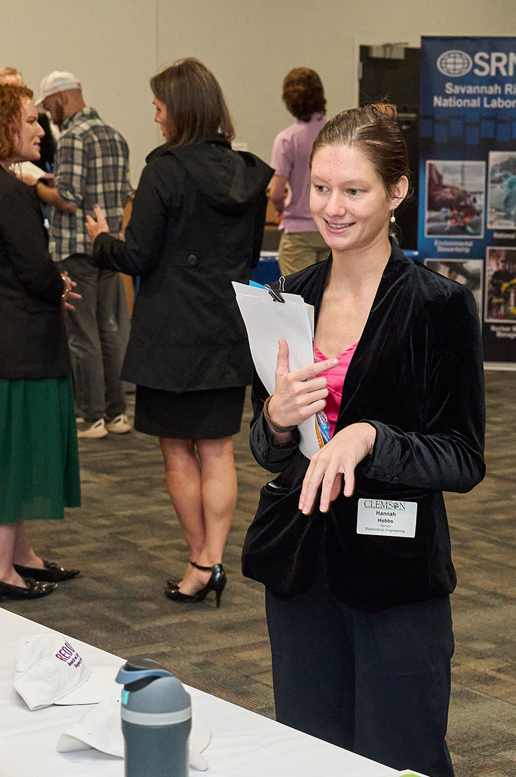 Woman holding papers and standing in an event space, talking.