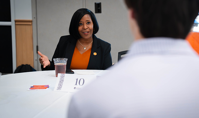 A female in a business suit speaks to a student from a seated position