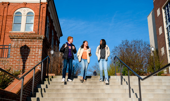 Three students walk down outdoor steps together on a sunny day, smiling and talking between brick academic buildings on Clemson's campus