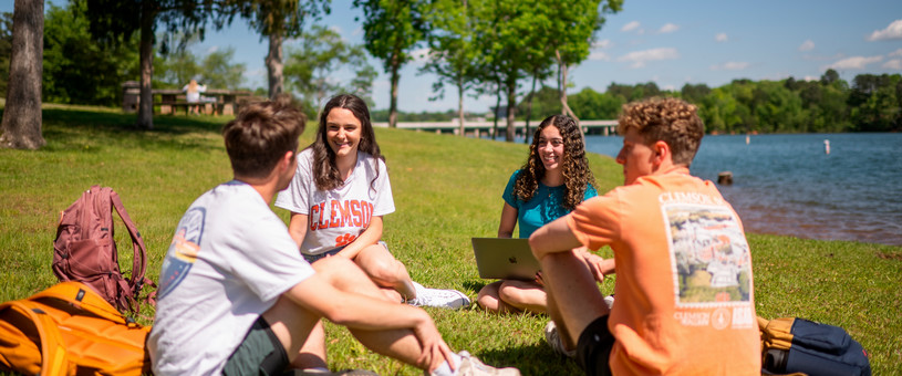 Four college students sit in a circle on a grassy lakeside lawn, talking and studying together on a sunny day. Backpacks and a laptop are visible, with trees, a dock, and calm water in the background. One student wears a Clemson shirt, and the group appears relaxed and engaged in conversation outdoors.