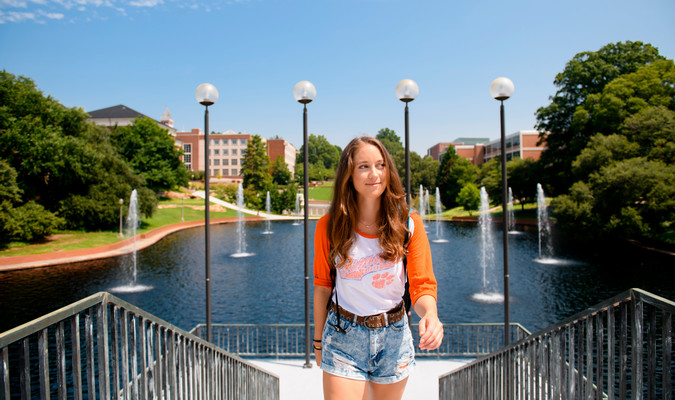Student walking up the steps on Clemson's Library Bridge on a sunny day with Clemson's Reflection Pond, trees, lampposts and academic buildings in the background.