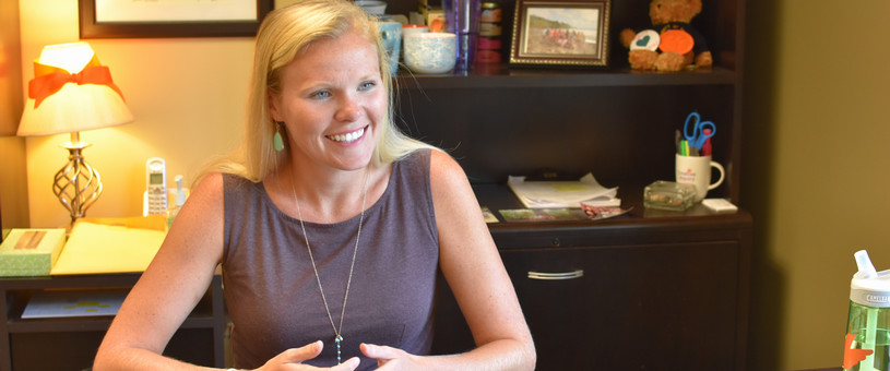 A woman sits at a desk in a professional office, smiling and engaged in conversation. A closed laptop and water bottle are on the desk, while a framed Clemson University diploma, family photos, office supplies, and decorative items are displayed on bookshelves behind her. Warm lighting creates a welcoming office environment.