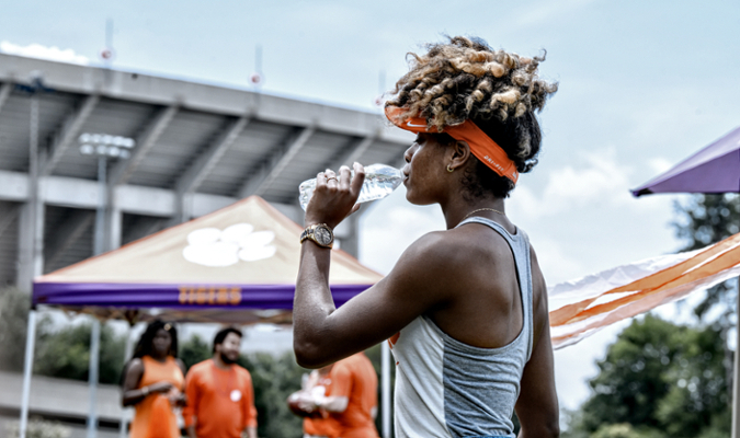 A female student drinks water at a Clemson football tailgate with Memorial Stadium in the background. 
