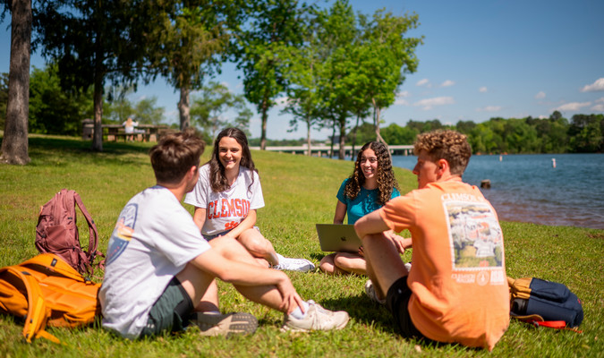 Four college students sit in a circle on a grassy lakeside lawn, talking and studying together on a sunny day. Backpacks and a laptop are visible, with trees, a dock, and calm water in the background. One student wears a Clemson shirt, and the group appears relaxed and engaged in conversation outdoors.