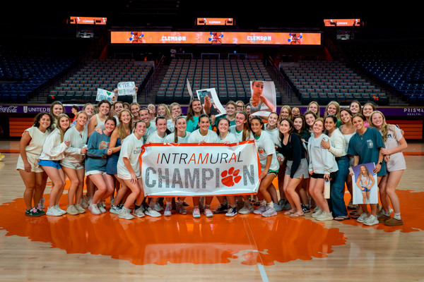 Large group of students on a basketball court holding an “Intramural Champions” banner and cheering with handmade signs in a Clemson arena.