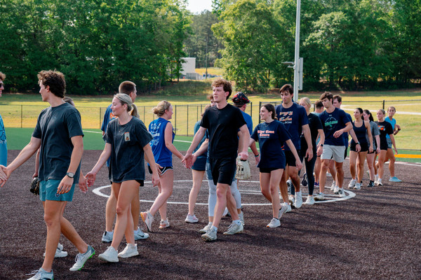 Group of young adults in athletic clothes walk across a sports field in a handshake line after a game.