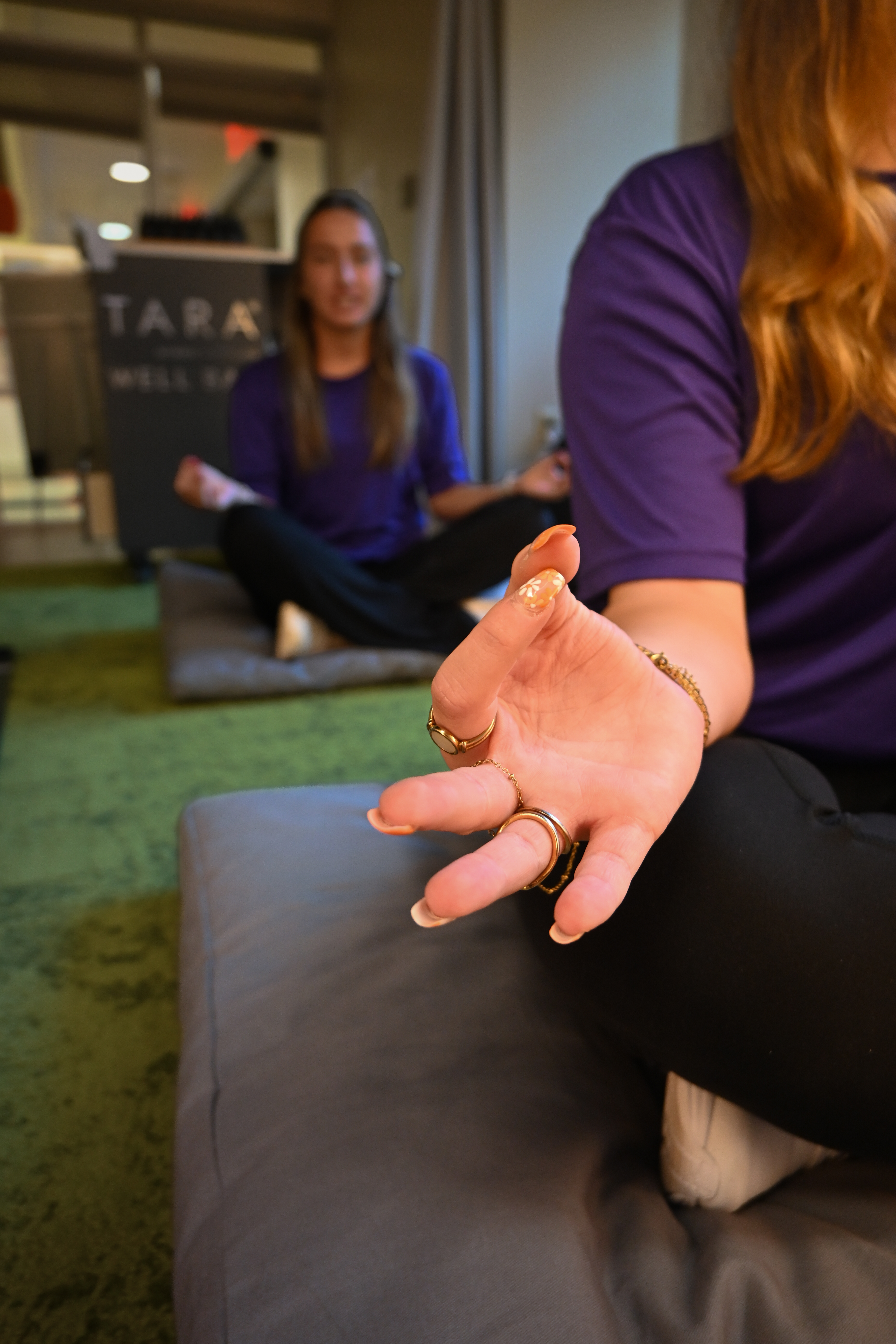 Person in a purple shirt holds a relaxed meditation hand pose in the foreground, with another person sitting cross-legged in the background.