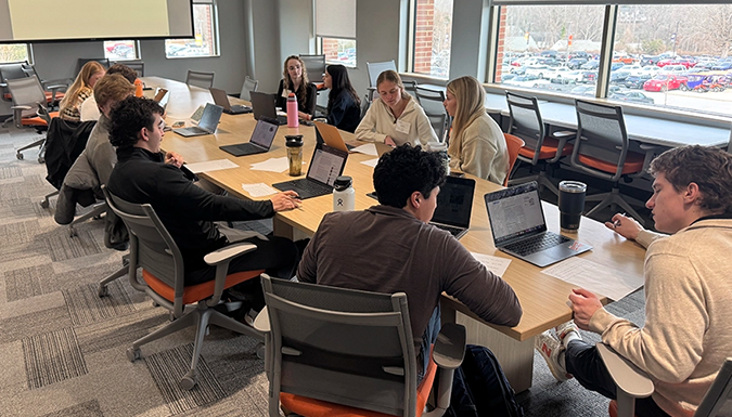 Students collaborate around a long table with laptops and papers in a classroom, with large windows overlooking a parking lot.