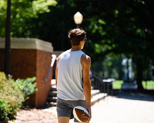 A young man carries a football while walking on campus.