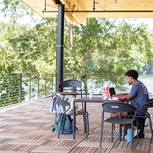 A student sits outside to study on their laptop on a covered deck near the lake