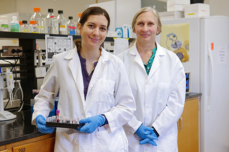 A student and professor wearing lab coats stand side by side in a science lab.