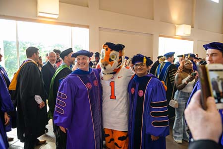 Two doctoral students pose with the Clemson Tiger mascot in their caps and gowns.