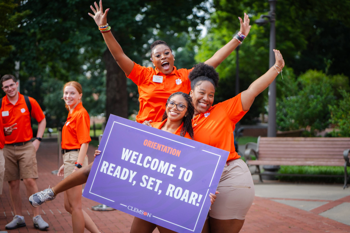 Three orientation ambassadors in orange polos hold a sign reading welcome to Ready, Set, Roar!