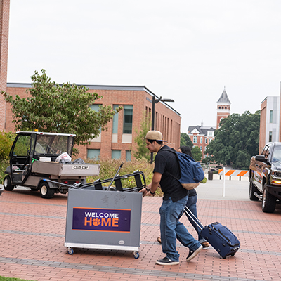 Students moving luggage and a cart labeled "WELCOME HOME" across a campus pathway.