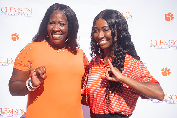 A mother and daughter smiling and posing together, wearing Clemson University apparel, with a backdrop featuring the university's logo.