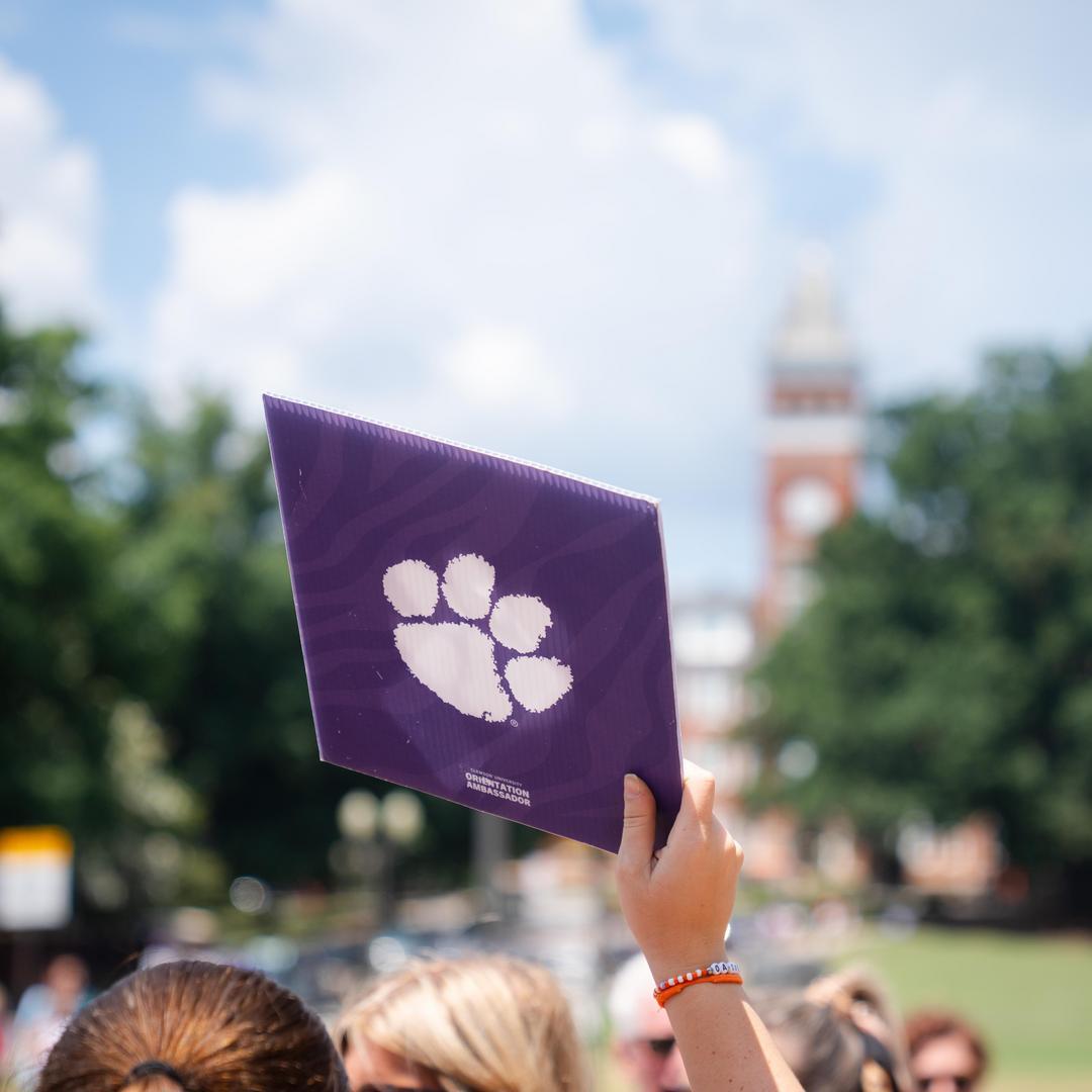 Hand holding a purple folder with a white paw print logo, with a campus clock tower blurred in the background.