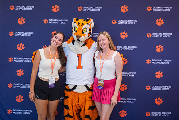 Two students pose for a photo with a tiger mascot in front of a backdrop featuring tiger paw and engineering and computing graphics.