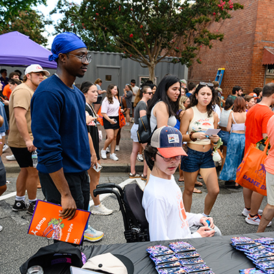 A diverse group of people at an outdoor event, with two individuals in focus: one sitting in a wheelchair and another standing nearby, observing the crowd.