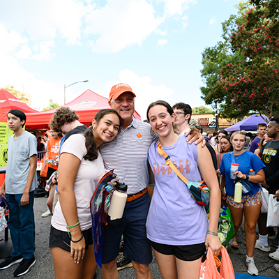 A joyful group photo of two young women posing with the Clemson President in an orange polo shirt at a lively outdoor event.