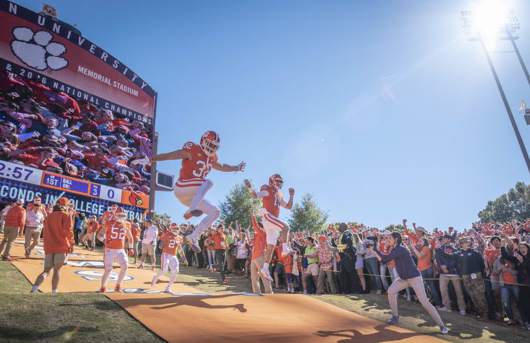 Clemson football players and coaches run down the hill at the start of a game with fans behind them.